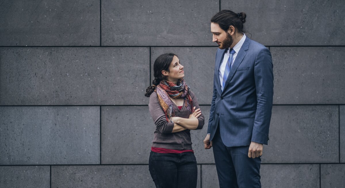 Mann und Frau sehen sich leicht lächelnd, aber etwas skeptisch an, sie ist sehr leger gekleidet, er trägt einen Anzug. Die beiden stehen vor einer Betonwand.