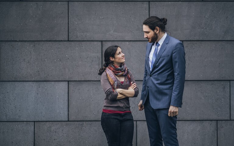 Mann und Frau sehen sich leicht lächelnd, aber etwas skeptisch an, sie ist sehr leger gekleidet, er trägt einen Anzug. Die beiden stehen vor einer Betonwand. | © Getty Images / Westend61