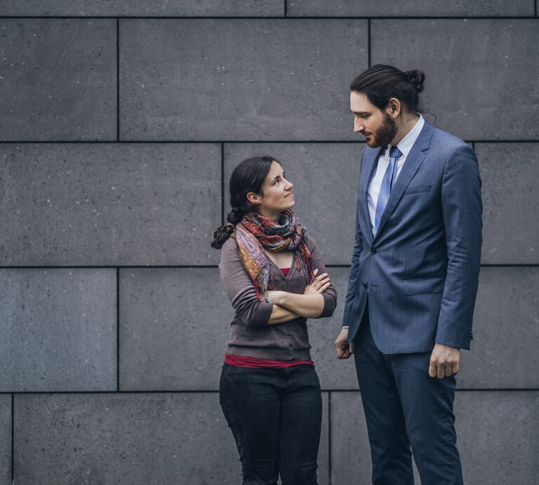 Mann und Frau sehen sich leicht lächelnd, aber etwas skeptisch an, sie ist sehr leger gekleidet, er trägt einen Anzug. Die beiden stehen vor einer Betonwand. | © Getty Images / Westend61
