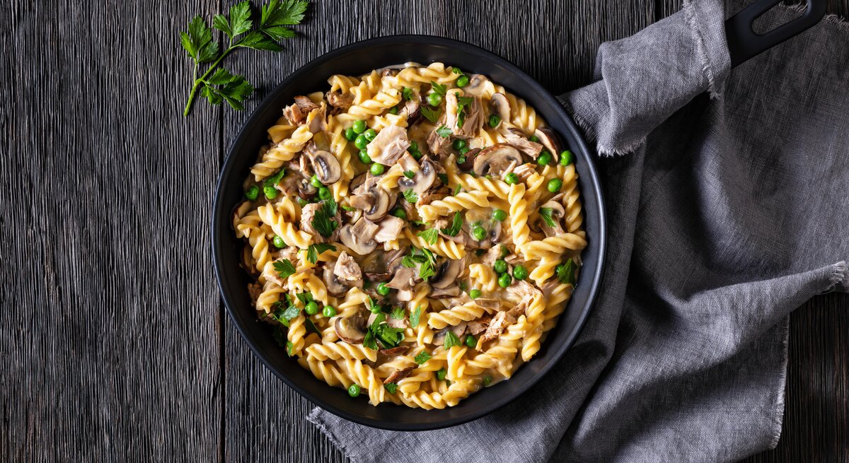 Thunfisch-Nudelpfanne mit Pilzen und Erbsen in Pfanne auf dunklem Untergrund. 