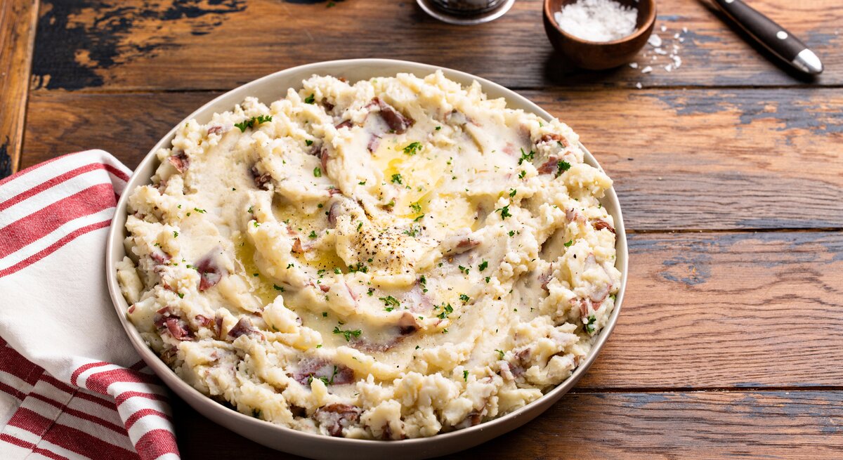 Frontalsicht: eine große Schale mit einem Knoblauch-Kartoffelpüree aus roten Kartoffeln steht auf einem Untergrund aus Holz. Daneben liegen ein Handtuch, Besteck, eine Knoblauchzehe und ein Schälchen mit Salz.