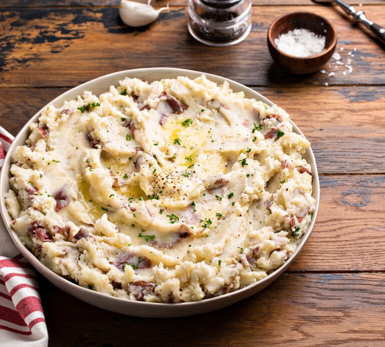 Frontalsicht: eine große Schale mit einem Knoblauch-Kartoffelpüree aus roten Kartoffeln steht auf einem Untergrund aus Holz. Daneben liegen ein Handtuch, Besteck, eine Knoblauchzehe und ein Schälchen mit Salz. | © Getty Images / VeselovaElena
