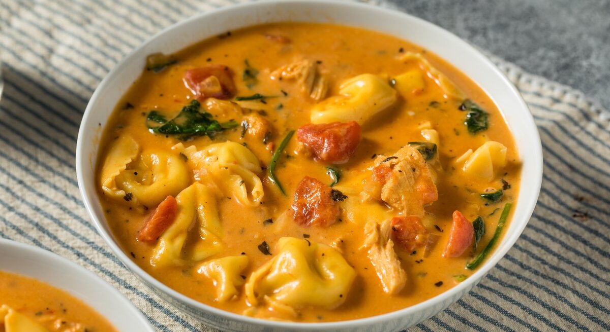 Ein weißer Teller mit der Tortellini-Suppe auf einem grauen Untergrund mit einem blau-weiß-gestreiften Tuch