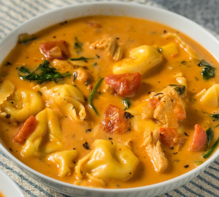 Ein weißer Teller mit der Tortellini-Suppe auf einem grauen Untergrund mit einem blau-weiß-gestreiften Tuch | © Shutterstock/ Brent Hofacker