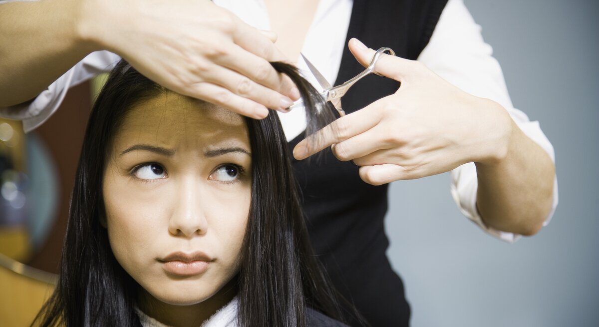 Junge Frau, die scheinbar besorgt schaut, während ihre Haare geschnitten werden. Die Friseurin, deren Gesicht nicht zu sehen ist, hält eine Haarsträhne der Frau und eine Schere bereit zum Schneiden. Die Frau trägt einen Friseurumhang und blickt 