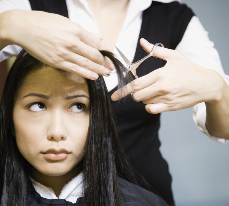 Junge Frau, die scheinbar besorgt schaut, während ihre Haare geschnitten werden. Die Friseurin, deren Gesicht nicht zu sehen ist, hält eine Haarsträhne der Frau und eine Schere bereit zum Schneiden. Die Frau trägt einen Friseurumhang und blickt  | © Getty Images/Andersen Ross Photography Inc