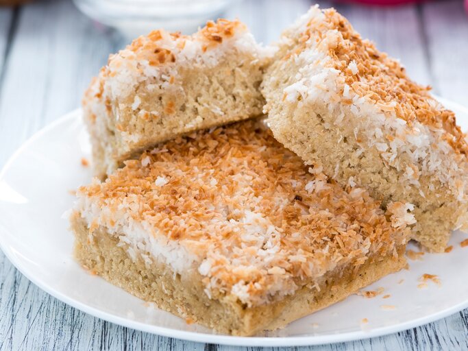 Drei Stücke vom saftigen Kokoskuchen auf einem weißen Teller und einem weißen Holztisch seitlich fotografiert. Im Hintergrund unscharf Backhandschuhe und eine halbe Kokosnuss. | © Shutterstock/ HandmadePictures