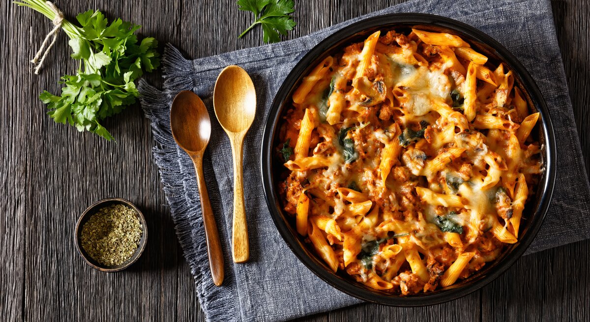 Gebackene Hähnchen-Nudeln mit Spinat und Pilzen in Tomatensoße auf schwarzem Teller. Dieser steht auf einem mit einem Geschirrtuch bedeckten Holztisch. Daneben liegen Holzlöffel, Petersilie und eine schale schwarzer, gemahlener Pfeffer.