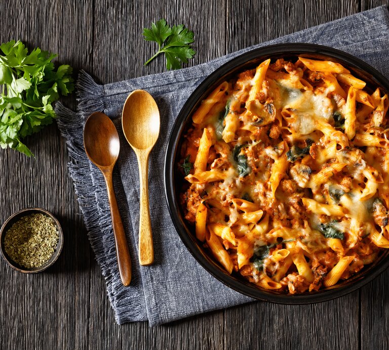 Gebackene Hähnchen-Nudeln mit Spinat und Pilzen in Tomatensoße auf schwarzem Teller. Dieser steht auf einem mit einem Geschirrtuch bedeckten Holztisch. Daneben liegen Holzlöffel, Petersilie und eine schale schwarzer, gemahlener Pfeffer. | © Getty Images/ 	from_my_point_of_view