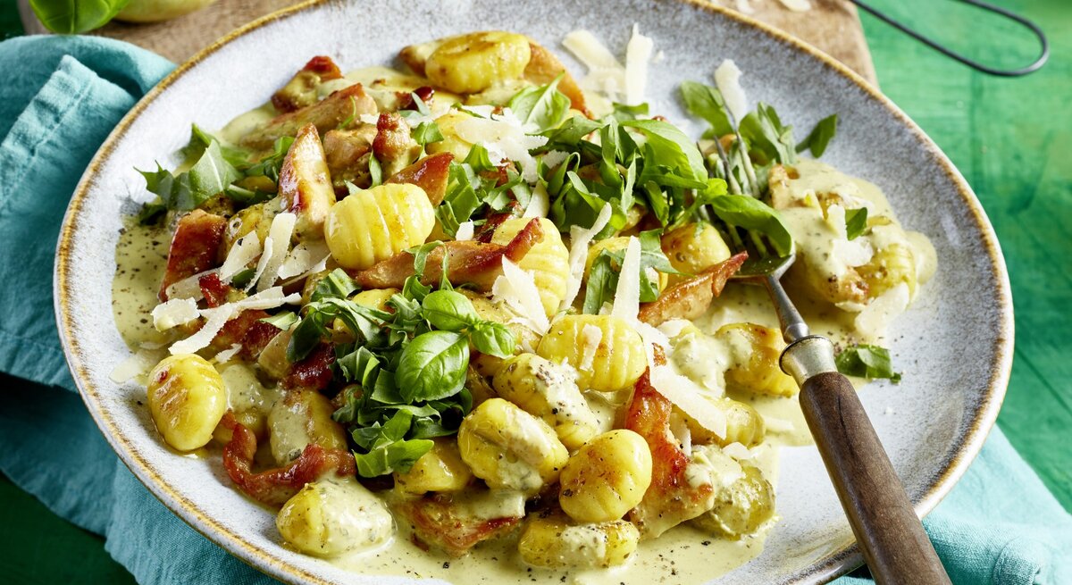 Pesto-Geschnetzeltes mit Gnocci, frischem Rucola und Basilikum und geriebenem Parmesan, serviert auf einem tiefen Keramikteller auf grünem Untergrund. 