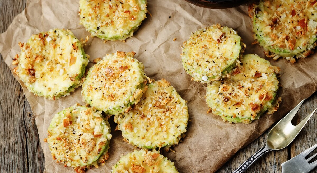 Zucchini-Parmesan-Chips auf einem Backpapier. Daneben Besteck und ein Ausschnitt einer Schale.