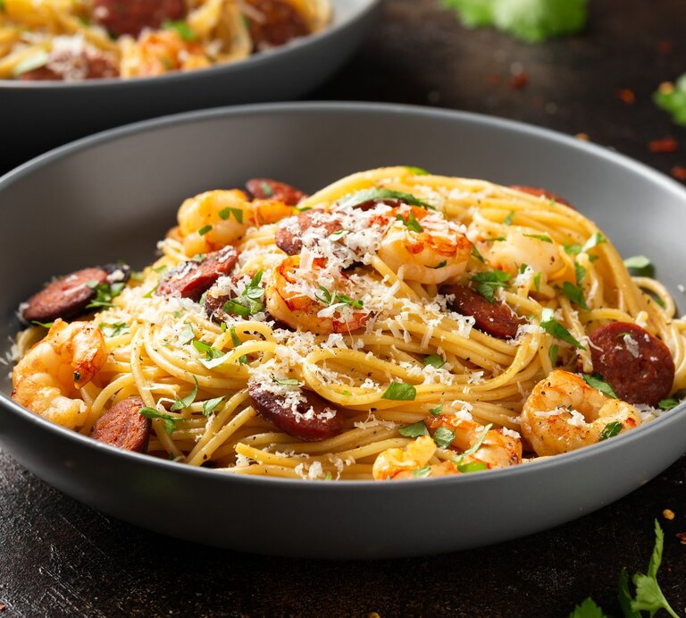 Ein grauer Teller mit der Garnelen- und Chorizopasta auf einem dunklen Holztisch mit vielen Kräutern und Käse bestreut. | © Shutterstock/ DronG