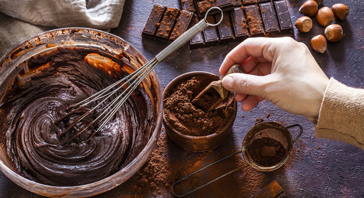 Dunkler Untergrund bedeckt mit allerlei Zutaten für einen Schokoladenkuchen, dessen Teig gerade in einer Schüssel angerührt wird. 