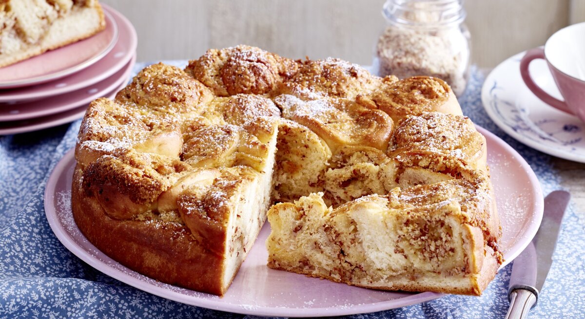 Ein Dänischer Butterkuchen steht auf einem Tisch. Daneben liegen mehrere Teller, ein Messer sowie eine Tasse mit einem Unterteller.