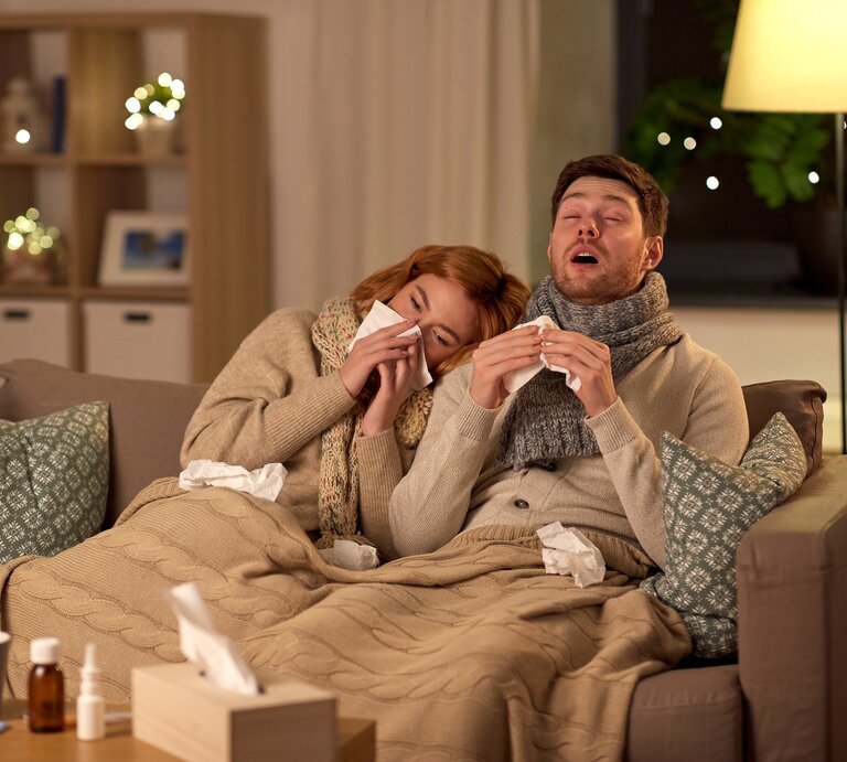 Ein junges Paar sitzt abends erkältet auf dem Sofa, er niest gerade, während sie sich an ihn angelehnt die Nase putzt. Beide sind in Schals, Strickjacken und Decken eingewickelt.  | © Getty Images/dolgachov