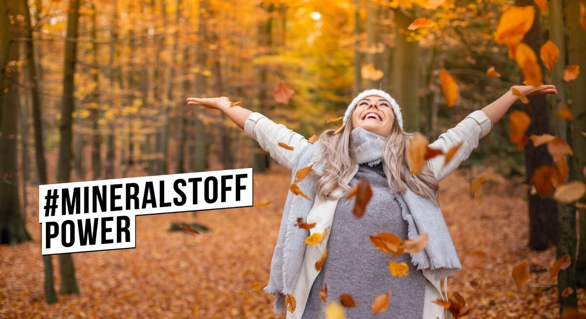 Eine glücklich lächelnde Frau mit grauem Hut und Schal streckt die Arme aus und genießt den Herbst im Wald. Sie ist umgeben von fallenden Blättern und einem Hintergrund aus leuchtend orangefarbenen Bäumen. Die Frau trägt eine graue Strickjacke und scheint den Moment der Ruhe in der Natur zu genießen. Über ihr ist das Hashtag #MINERALSTOFFPOWER in schwarzer Schrift auf einem weißen Balken abgebildet.