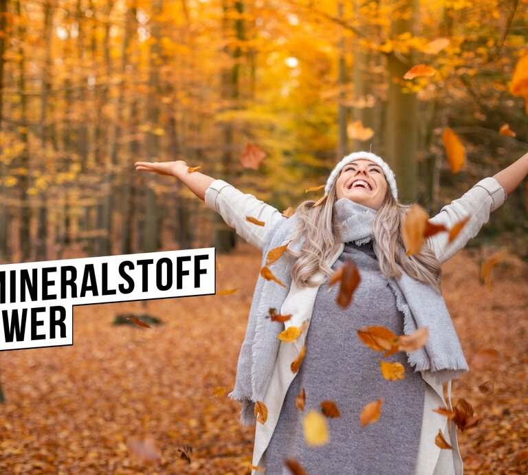 Eine glücklich lächelnde Frau mit grauem Hut und Schal streckt die Arme aus und genießt den Herbst im Wald. Sie ist umgeben von fallenden Blättern und einem Hintergrund aus leuchtend orangefarbenen Bäumen. Die Frau trägt eine graue Strickjacke und scheint den Moment der Ruhe in der Natur zu genießen. Über ihr ist das Hashtag #MINERALSTOFFPOWER in schwarzer Schrift auf einem weißen Balken abgebildet. | © iStock.com/Penelope Graßhoff