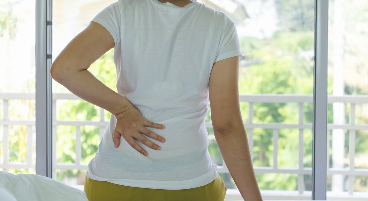 Frau sitzt auf dem Bett und legt eine Hand auf den Rücken, da sie Scherzen aufgrund einer Nierenbeckenentzündung hat.