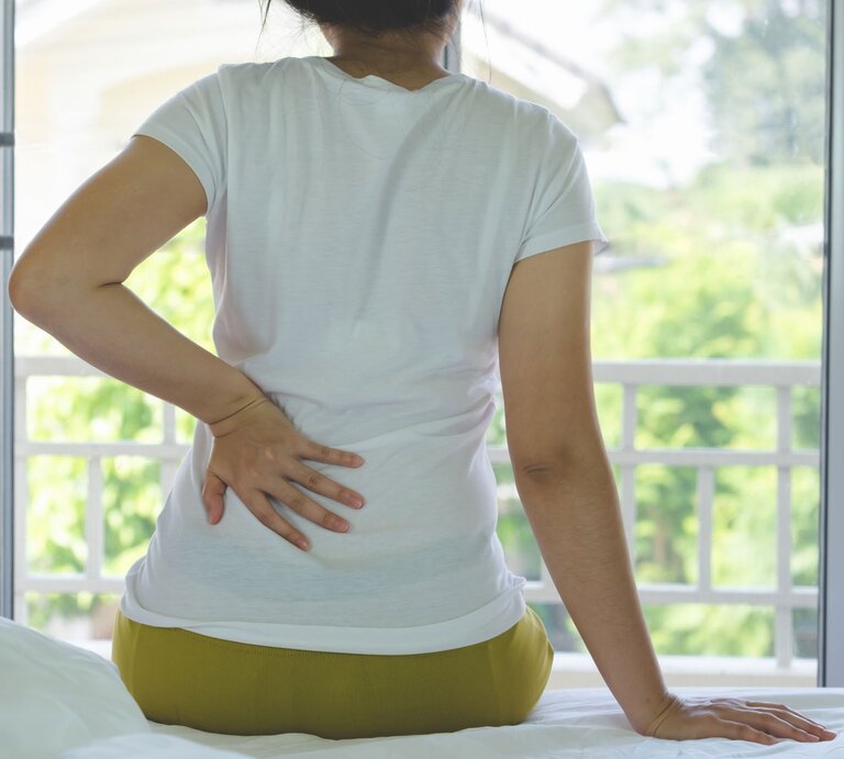 Frau sitzt auf dem Bett und legt eine Hand auf den Rücken, da sie Scherzen aufgrund einer Nierenbeckenentzündung hat. | © iStock/Iamstocker