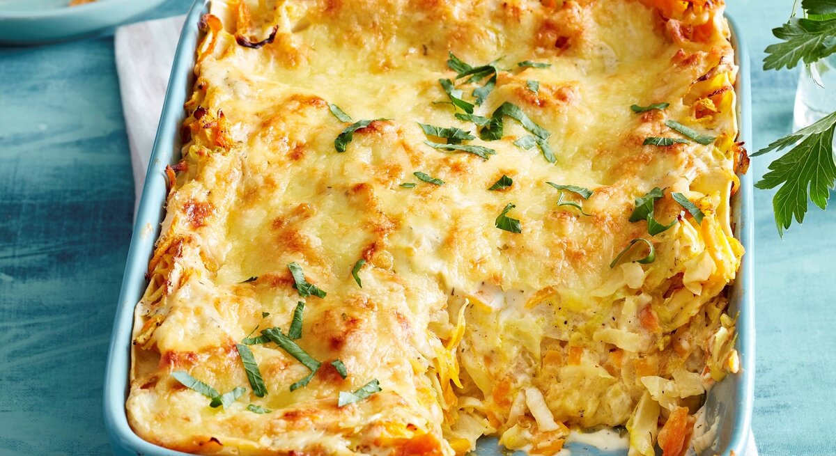 Blaue Ofenform mit angeschnittener Gemüse-Lasagne, Geschirr und Kräuter im blauen Hintergrund.