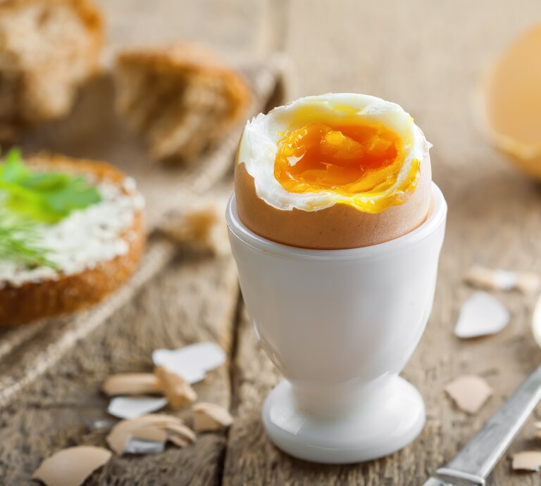 Geköpftes Frühstücksei in weißem Eierbecher auf Holz.  | © GettyImages / Derkien