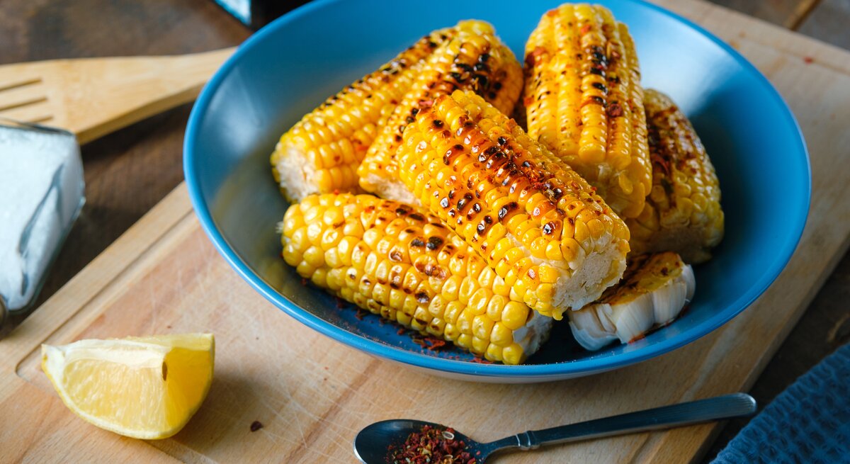 Das Bild zeigt eine Nahaufnahme von gegrillten Maiskolben, die appetitlich in einer tiefen blauen Schüssel serviert werden. Die Maiskörner haben eine leuchtend gelbe Farbe mit Grillspuren, die ihnen ein rustikales Aussehen verleihen. Neben der Schüssel liegt ein Stück Zitrone, dessen frische Farbe einen schönen Kontrast zum Gelb des Maises bildet. Auf dem Schneidebrett vor der Schüssel ist ein Löffel mit einer Gewürzmischung zu sehen, was darauf hindeutet, dass der Mais gewürzt wurde. Im Hintergrund sind Küchenutensilien und eine klare Flasche, möglicherweise Öl oder Essig, angedeutet, was die häusliche und gemütliche Atmosphäre unterstreicht.