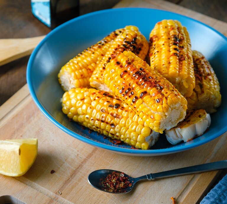 Das Bild zeigt eine Nahaufnahme von gegrillten Maiskolben, die appetitlich in einer tiefen blauen Schüssel serviert werden. Die Maiskörner haben eine leuchtend gelbe Farbe mit Grillspuren, die ihnen ein rustikales Aussehen verleihen. Neben der Schüssel liegt ein Stück Zitrone, dessen frische Farbe einen schönen Kontrast zum Gelb des Maises bildet. Auf dem Schneidebrett vor der Schüssel ist ein Löffel mit einer Gewürzmischung zu sehen, was darauf hindeutet, dass der Mais gewürzt wurde. Im Hintergrund sind Küchenutensilien und eine klare Flasche, möglicherweise Öl oder Essig, angedeutet, was die häusliche und gemütliche Atmosphäre unterstreicht. | © Getty Images / Aleksandr Zubkov