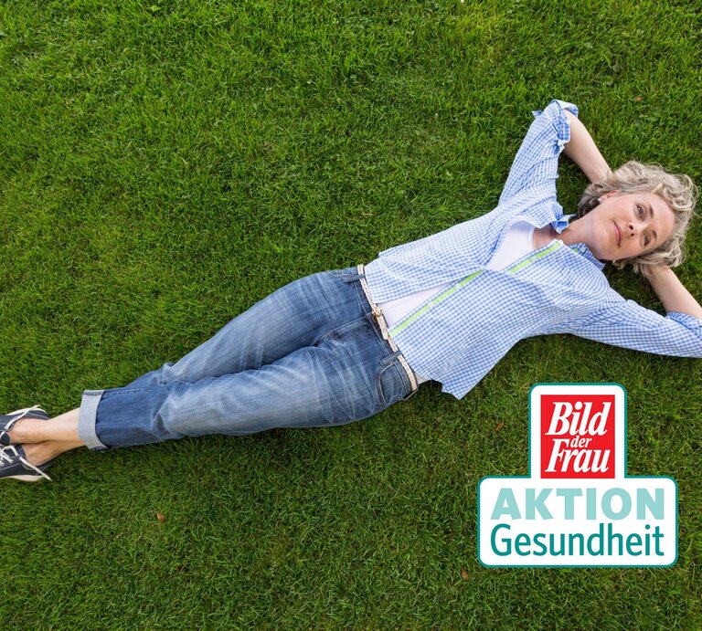 Eine entspannte reife Frau mit lockigen grauen Haaren liegt zurückgelehnt auf einer saftig grünen Wiese. Sie trägt eine blaue karierte Bluse, blaue Jeans und schwarze Sneakers. Ihre Arme sind hinter dem Kopf verschränkt, was ein Gefühl von Ruhe und Entspannung vermittelt. In der unteren rechten Ecke des Bildes befindet sich ein Logo mit der Aufschrift "Bild der Frau AKTION Gesundheit". | © iStock/solarisimages [M]