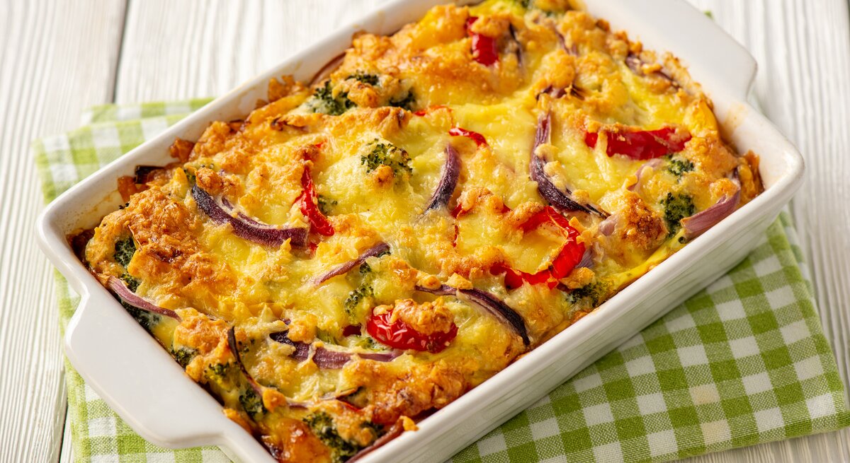 Weiße Auflaufform gefüllt mit Gemüse-Kartoffelauflauf mit Brokkoli und Paprika. Die Form ist platziert auf einem grün-weißem Geschirrtuch. 