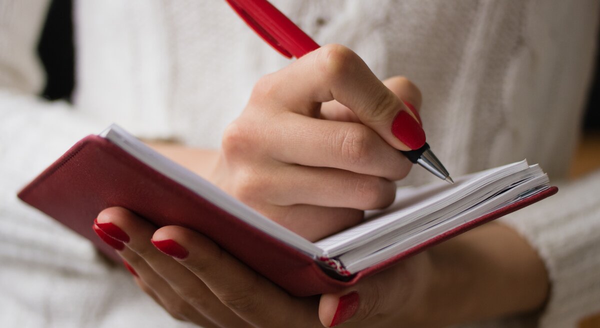 Frau mit roten Fingernägeln schreibt mit einem Kugelschreiber handschriftlich in ein Notizbuch. 