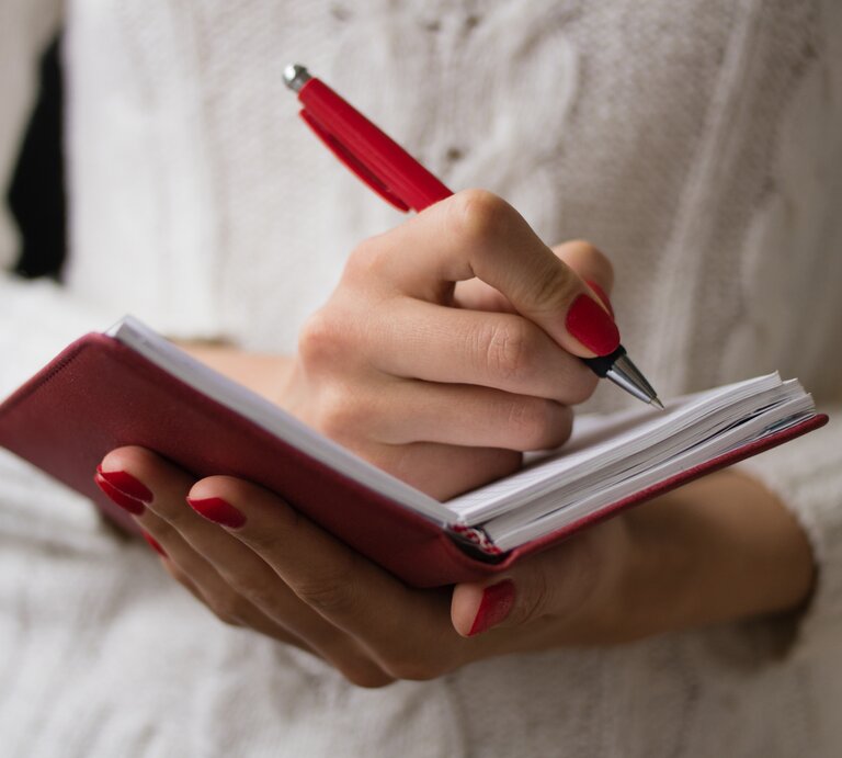 Frau mit roten Fingernägeln schreibt mit einem Kugelschreiber handschriftlich in ein Notizbuch.  | © iStock/iprogressman