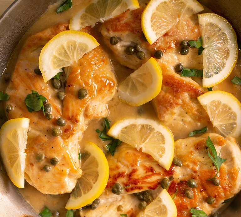 Eine Pfanne mit dem Hähnchen, Kapern und der Zitrone auf einem hellen Holztisch. | © Shutterstock/ Catherine Murray