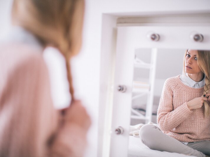 Frau, die vor einem Spiegel sitzt und sich selbst dabei betrachtet, wie sie ihr blondes Haar zu einem Zopf flicht. Sie trägt einen gemütlichen rosafarbenen Pullover und darunter ein hellblaues Hemd | © iStock/AleksandarNakic