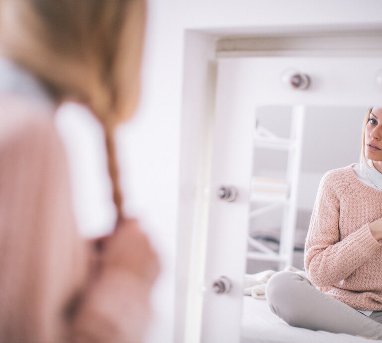 Frau, die vor einem Spiegel sitzt und sich selbst dabei betrachtet, wie sie ihr blondes Haar zu einem Zopf flicht. Sie trägt einen gemütlichen rosafarbenen Pullover und darunter ein hellblaues Hemd | © iStock/AleksandarNakic