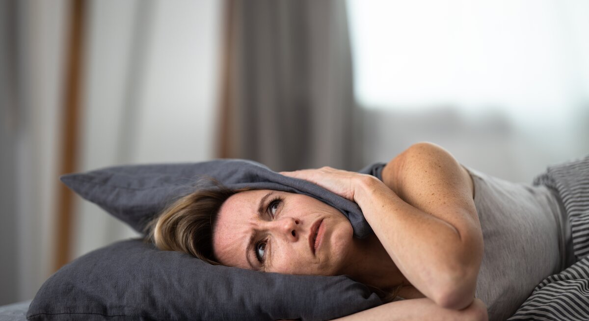 Eine besorgte mittelalterliche Frau liegt im Bett und stützt ihren Kopf auf einem Kissen ab, während sie das Kissen mit beiden Händen umklammert. Ihr Gesicht zeigt Anzeichen von Stress oder Schlaflosigkeit, mit sichtbaren Falten und einem müden Ausdruck. Der Hintergrund ist unscharf, was den Fokus auf ihre besorgte Miene lenkt. Das Bild könnte im Kontext von Schlafstörungen, Stressbewältigung oder gesundheitlichen Auswirkungen von Schlafmangel verwendet werden.