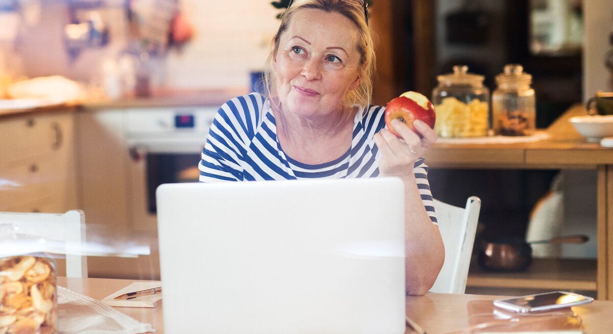 Reife Frau sitzt an einem Laptop und hat einen Apfel in der Hand. Sie sieht sehr zuversichtlich aus. 