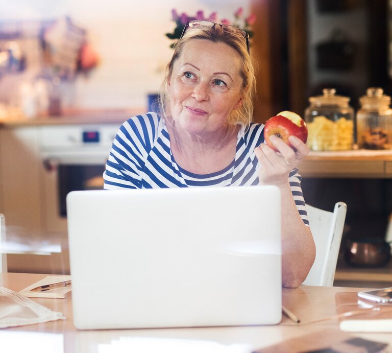 Reife Frau sitzt an einem Laptop und hat einen Apfel in der Hand. Sie sieht sehr zuversichtlich aus.  | © iStock/GrapeImages