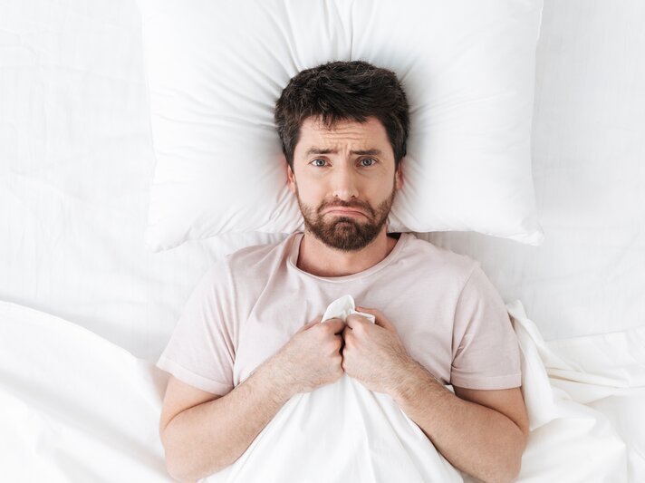 Mann mittleren Alters, der im Bett liegt und die Bettdecke bis zur Brust hochgezogen hat. Sein Gesichtsausdruck wirkt besorgt oder ängstlich, er hält die Decke mit beiden Händen fest. Er trägt ein einfaches, hellfarbenes T-Shirt und liegt auf einem weißen Kissen. | © Shutterstock/Dean Drobot