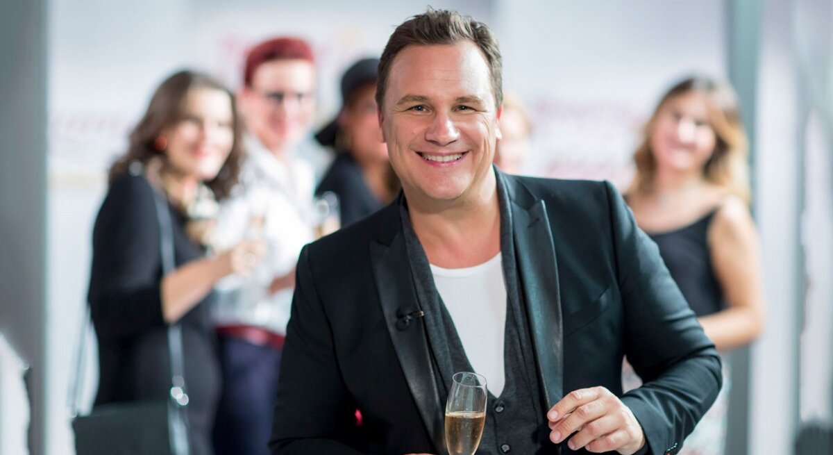 Ein lächelnder Guido Kretschmer in einem schwarzen Blazer und weißem T-Shirt hält ein Glas Sekt und blickt in die Kamera. Er steht im Vordergrund, während im Hintergrund unscharf weitere Personen zu sehen sind, die ebenfalls Getränke halten und sich in einer offensichtlich geselligen Atmosphäre befinden.