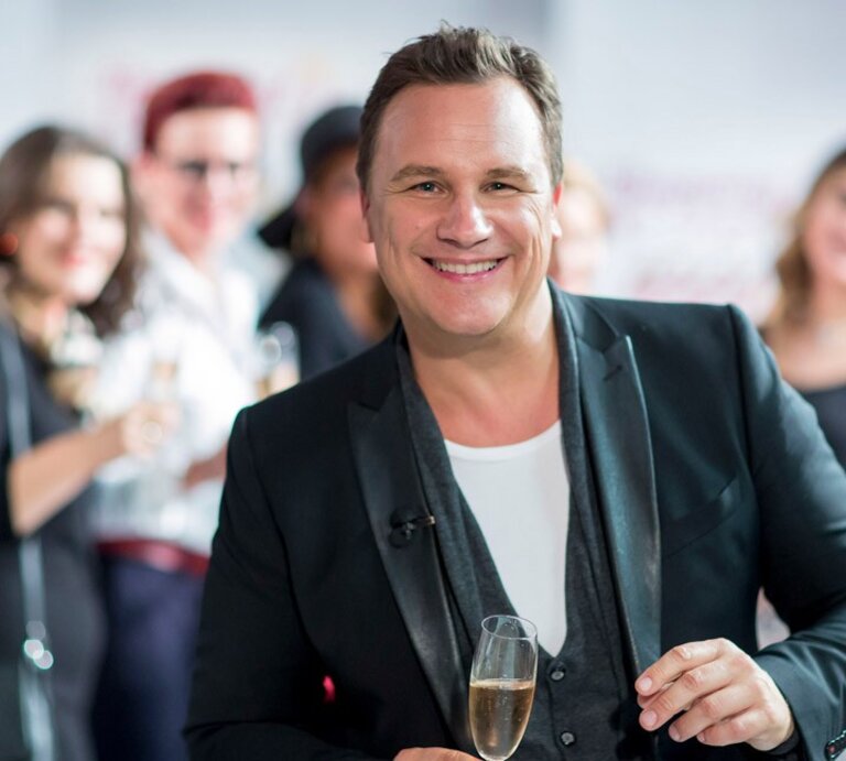 Ein lächelnder Guido Kretschmer in einem schwarzen Blazer und weißem T-Shirt hält ein Glas Sekt und blickt in die Kamera. Er steht im Vordergrund, während im Hintergrund unscharf weitere Personen zu sehen sind, die ebenfalls Getränke halten und sich in einer offensichtlich geselligen Atmosphäre befinden. | © VOX