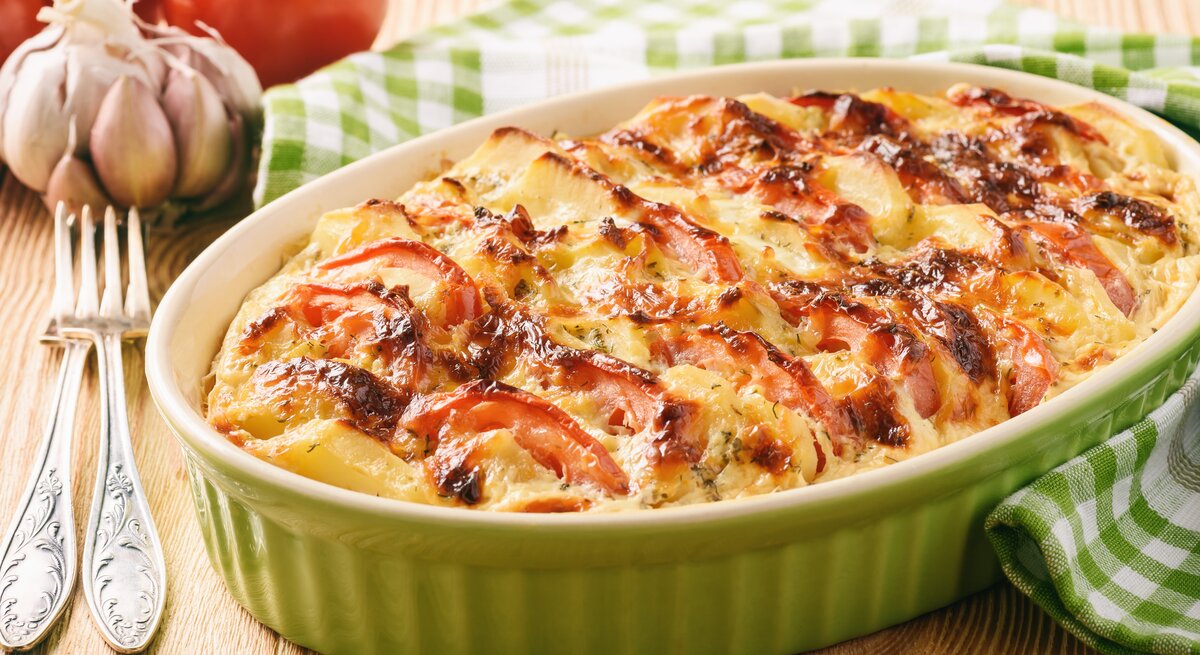 Eine Auflaufform mit Kürbis-Kartoffel-Auflauf mit Tomaten und Käse überbacken, daneben Tomaten, Knoblauch und eine Gabel.