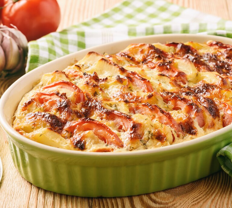 Eine Auflaufform mit Kürbis-Kartoffel-Auflauf mit Tomaten und Käse überbacken, daneben Tomaten, Knoblauch und eine Gabel. | © Adobe Stock/ O.B.