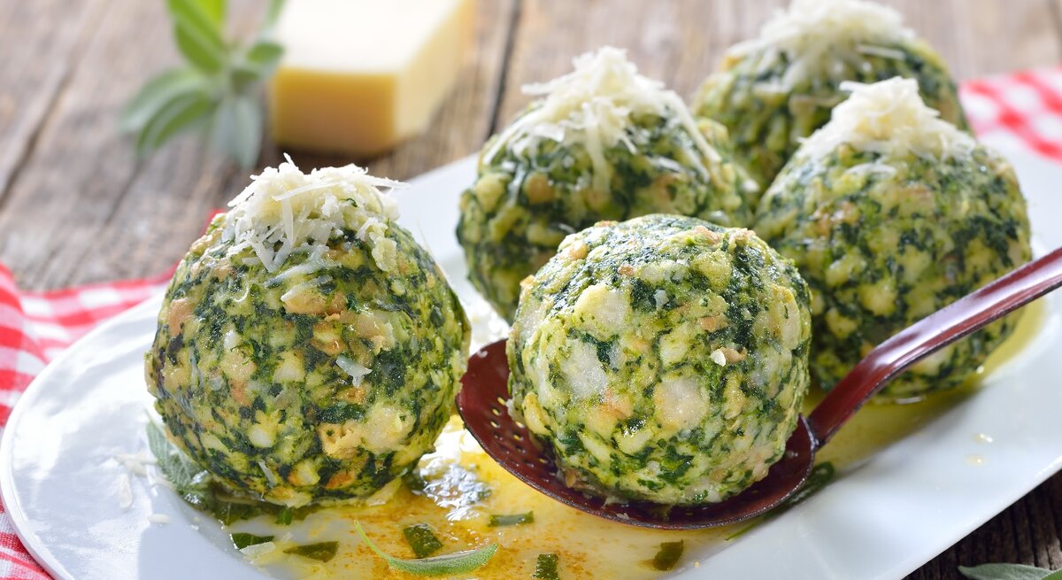 Fünf Spinatknödel auf einem länglichen weißen Teller mit der Salbeibutter. Im Hintergrund ein rot-weiß-kariertes Tuch und ein Stück ganzer Parmesan.