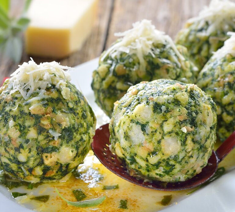 Fünf Spinatknödel auf einem länglichen weißen Teller mit der Salbeibutter. Im Hintergrund ein rot-weiß-kariertes Tuch und ein Stück ganzer Parmesan. | © Shutterstock/ Karl Allgaeuer