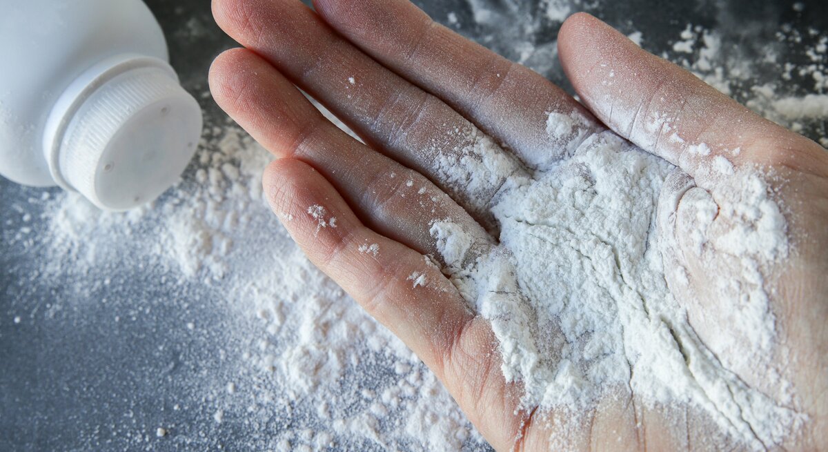  Hand, auf deren Fingern sich Babypuder befindet. Im Hintergrund ist eine umgestürzte Flasche mit Babypuder zu erkennen, aus der etwas Puder auf die Oberfläche gestreut wurde.