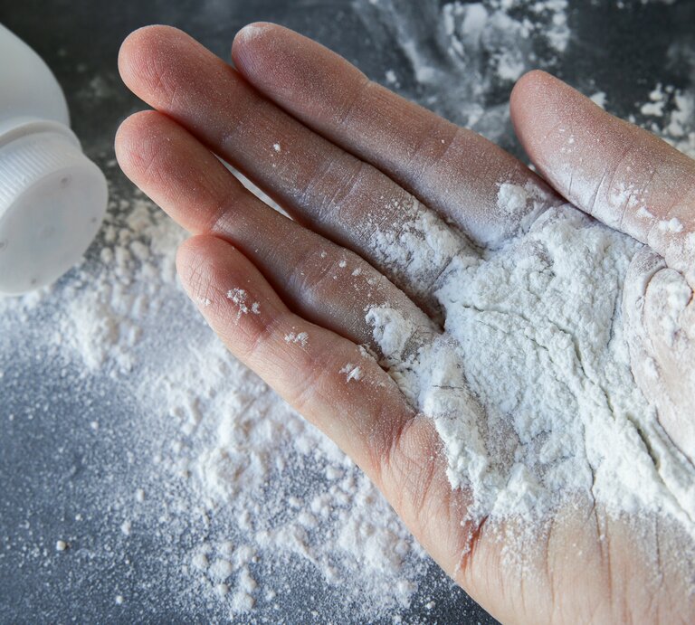  Hand, auf deren Fingern sich Babypuder befindet. Im Hintergrund ist eine umgestürzte Flasche mit Babypuder zu erkennen, aus der etwas Puder auf die Oberfläche gestreut wurde. | © iStock/ThitareeSarmkasat