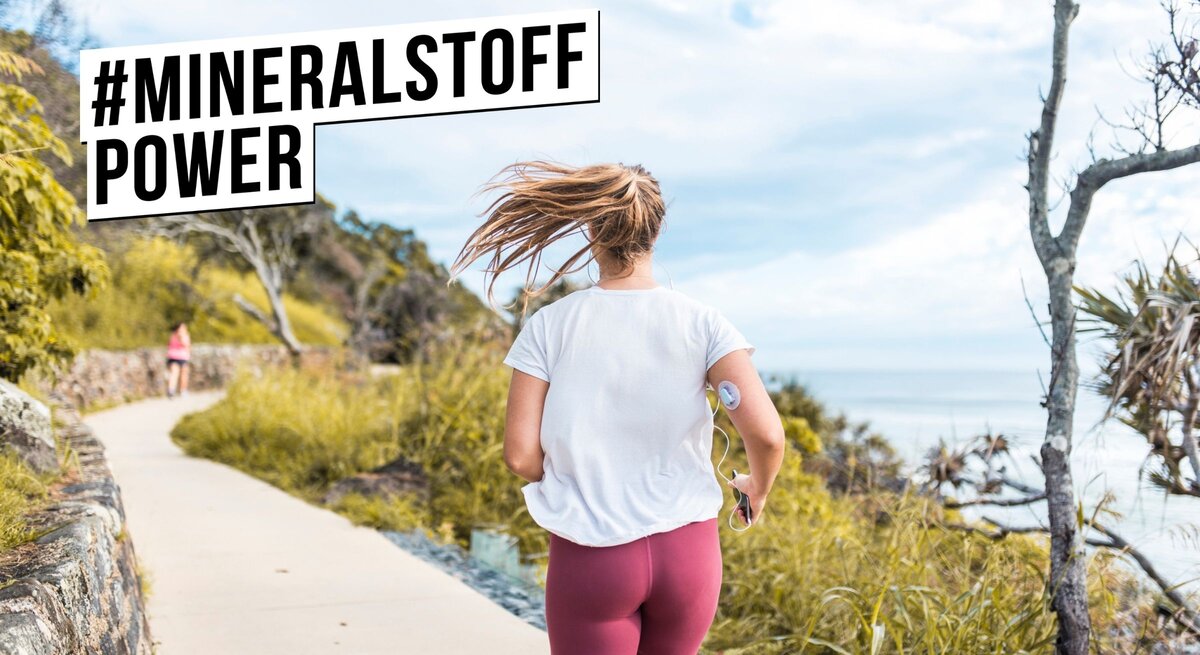 Eine aktive Frau in Freizeitkleidung joggend von hinten gesehen auf einem Küstenweg. Sie trägt ein weißes T-Shirt, rote Sportleggings und hat eine Fitnessuhr am Arm. Ihr Haar weht im Wind, während sie sich bewegt. Im Vordergrund ist das Hashtag #MINERALSTOFFPOWER in einem schwarzen Balken prominent dargestellt, was auf die Wichtigkeit von Mineralstoffen für einen gesunden Lebensstil und bei der Bewältigung von Gesundheitszuständen wie Diabetes hinweist. Die Szene strahlt Energie und Gesundheitsbewusstsein aus.