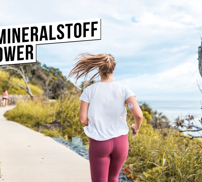 Eine aktive Frau in Freizeitkleidung joggend von hinten gesehen auf einem Küstenweg. Sie trägt ein weißes T-Shirt, rote Sportleggings und hat eine Fitnessuhr am Arm. Ihr Haar weht im Wind, während sie sich bewegt. Im Vordergrund ist das Hashtag #MINERALSTOFFPOWER in einem schwarzen Balken prominent dargestellt, was auf die Wichtigkeit von Mineralstoffen für einen gesunden Lebensstil und bei der Bewältigung von Gesundheitszuständen wie Diabetes hinweist. Die Szene strahlt Energie und Gesundheitsbewusstsein aus. | © iStock.com/AzmanJaka