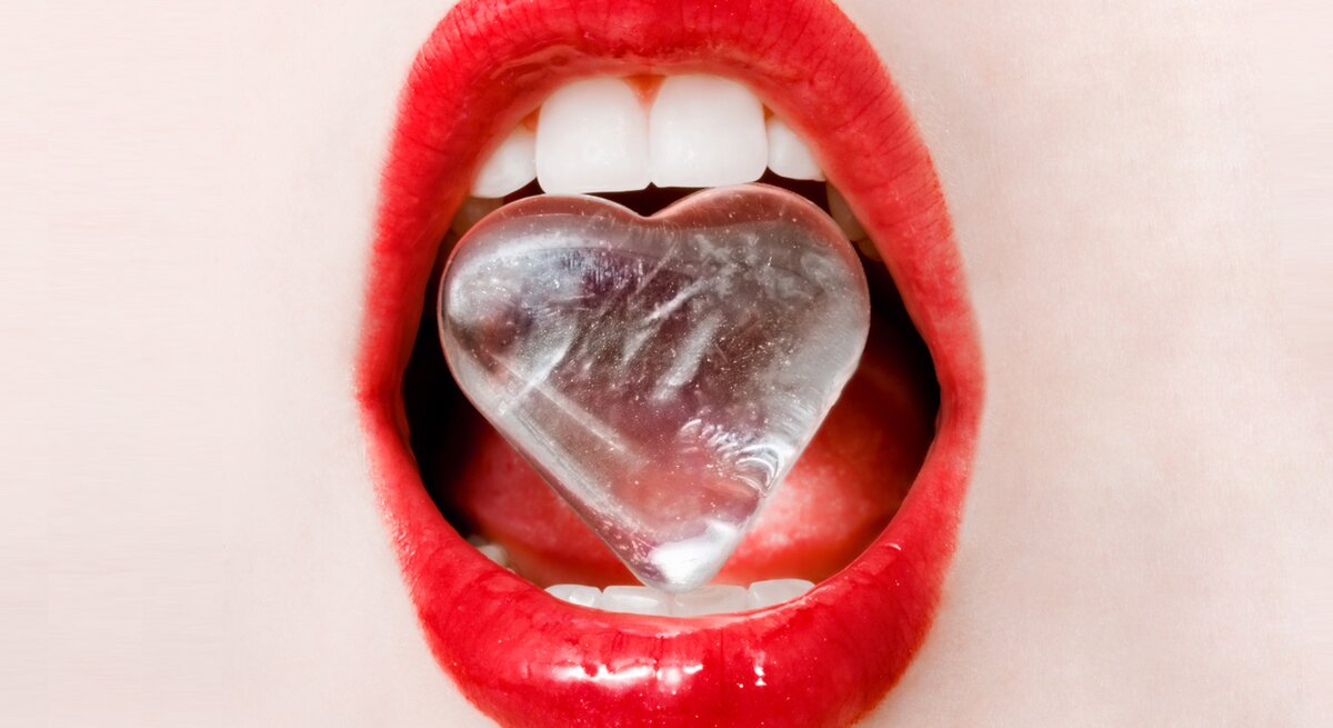 Close-up von roten Lippen. Die Zähne halten einen Eiswürfen in Herzform. 