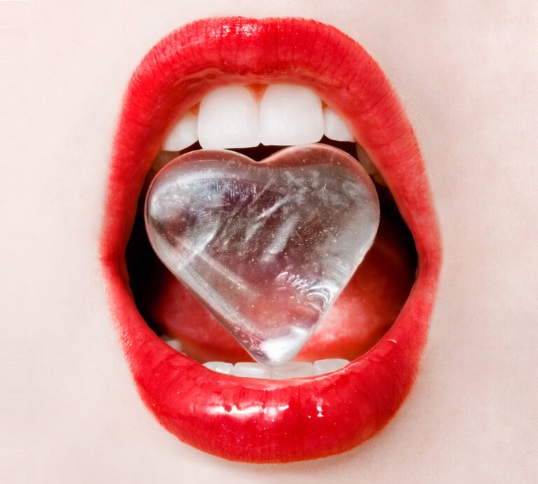 Close-up von roten Lippen. Die Zähne halten einen Eiswürfen in Herzform.  | © iStock/anneleven